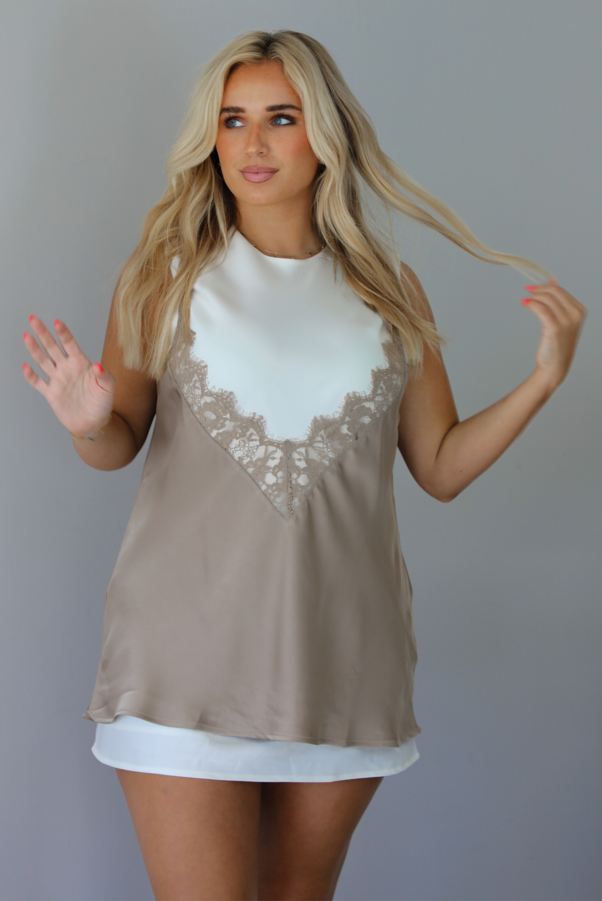 Woman wearing a taupe and white dress that looks like a lace tank overlayed over a white high neck mini dress, but it's all one piece indoors against a white wall.