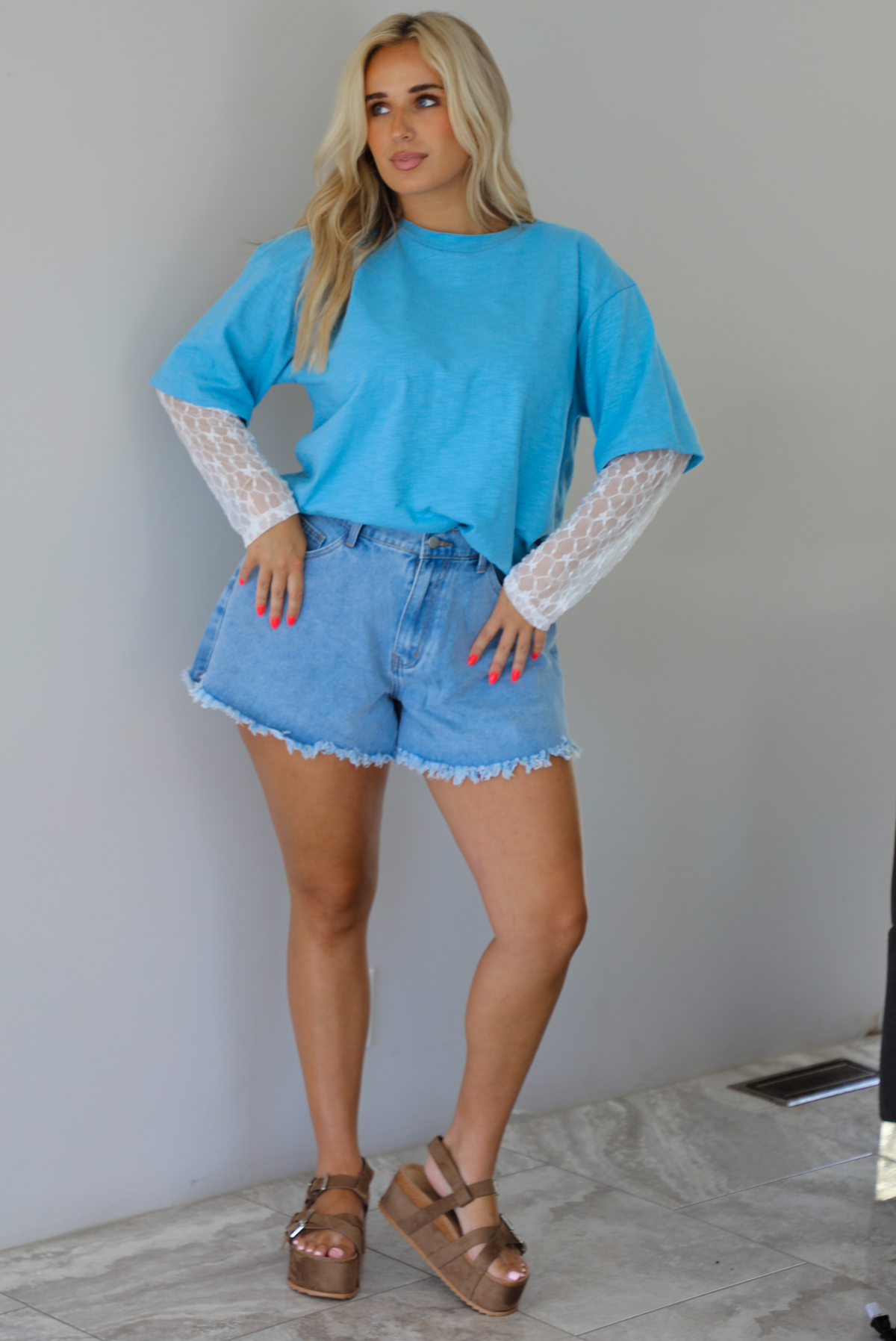 Woman wearing an oversized blue t-shirt with white lace long sleeves with medium wash denim shorts indoors against a white wall with white marble flooring. 
