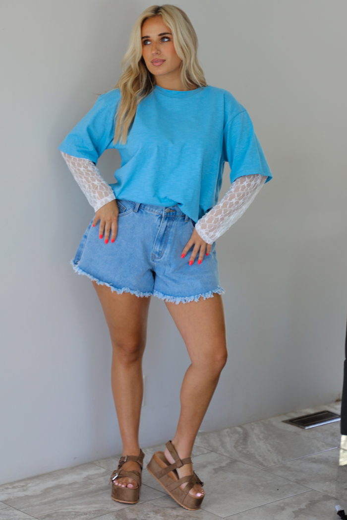 Woman wearing an oversized blue t-shirt with white lace long sleeves with medium wash denim shorts indoors against a white wall with white marble flooring. 