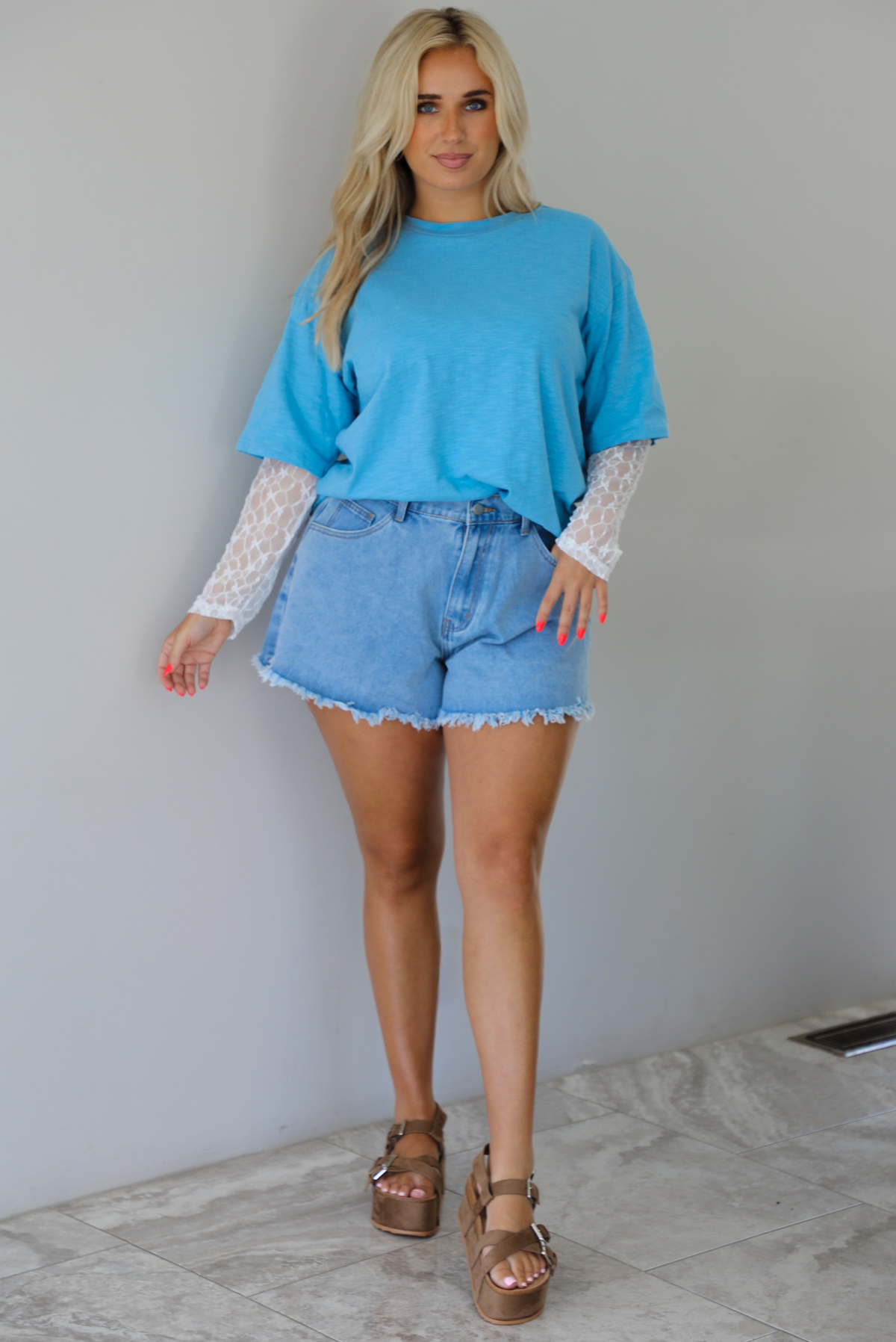 Woman wearing an oversized blue t-shirt with white lace long sleeves with medium wash denim shorts indoors against a white wall with white marble flooring. 