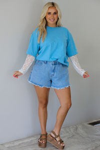 Woman wearing an oversized blue t-shirt with white lace long sleeves with medium wash denim shorts indoors against a white wall with white marble flooring. 