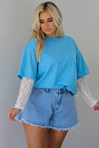 Woman wearing an oversized blue t-shirt with white lace long sleeves with medium wash denim shorts indoors against a white wall. The photo shows the tee tucked into the denim shorts to give it a cropped look. 