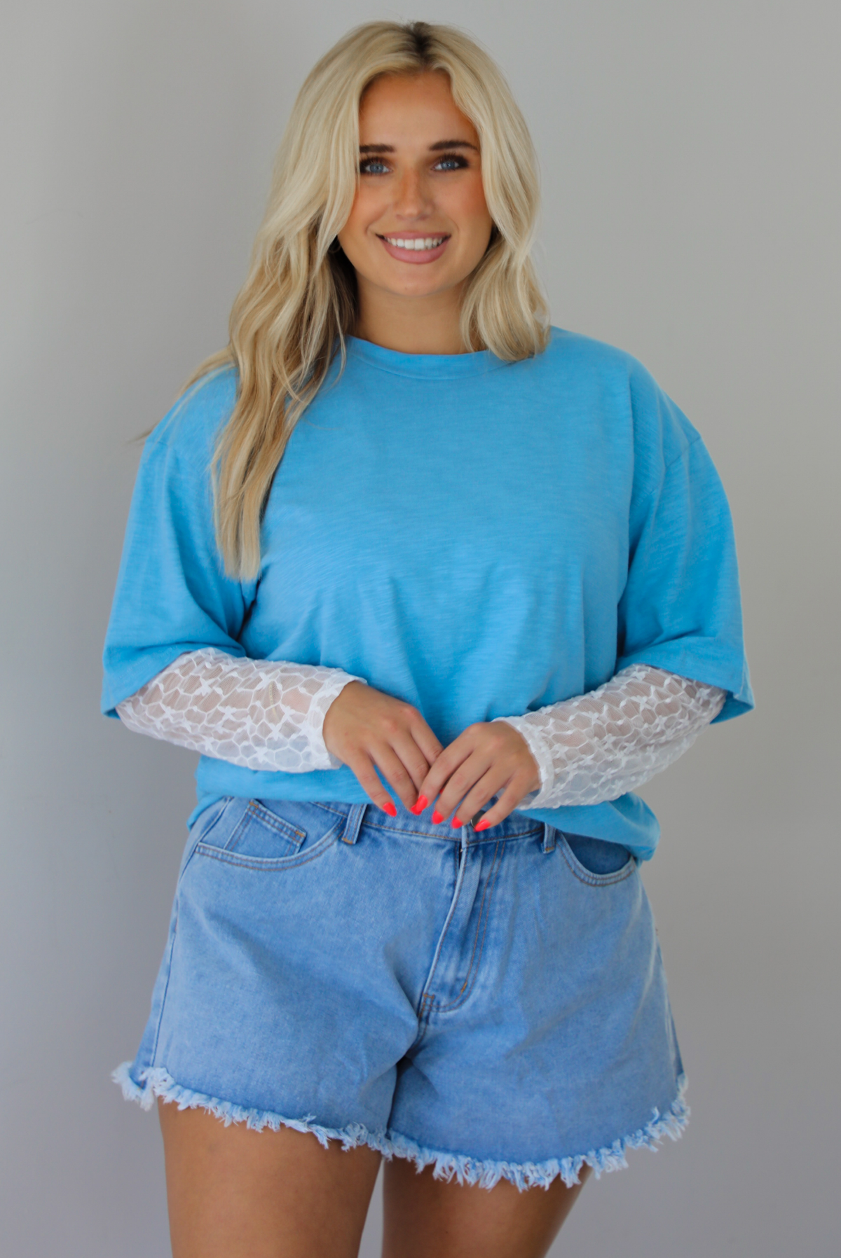 Woman wearing an oversized blue t-shirt with white lace long sleeves with medium wash denim shorts indoors against a white wall. The photo shows the tee tucked into the denim shorts to give it a cropped look. 