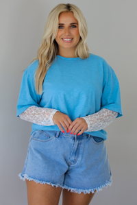 Woman wearing an oversized blue t-shirt with white lace long sleeves with medium wash denim shorts indoors against a white wall. The photo shows the tee tucked into the denim shorts to give it a cropped look. 