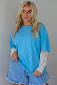 Woman wearing an oversized blue t-shirt with white lace long sleeves with medium wash denim shorts indoors against a white wall. The photo shows the tee untucked to display the full length.  