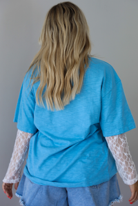 Woman wearing an oversized blue t-shirt with white lace long sleeves with medium wash denim shorts indoors against a white wall. The photo shows the back of the tee. 