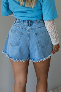 Woman wearing blue denim shorts that are high waisted with a raw edged hem indoors against a white wall. The photo shows the back of the shorts, featuring the elastic waistline and functional pockets in the back.