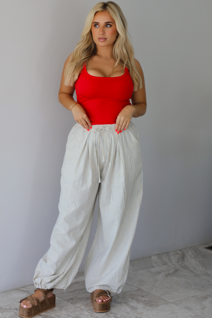 Woman wearing a red tank tucked info ecru lounge pants that have a blue striped design, high waistline, and functional pockets on either side indoors against a white wall with white marble flooring. 