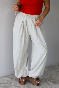 Woman wearing lounge pants that are an ecru color with a blue striped pattern, are high waisted with an elastic and drawstring waistline, and have a slouched design until they cinch at the ankles indoors against a white wall with white marble flooring. 
