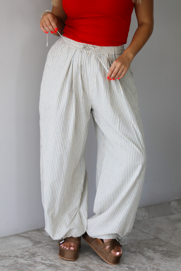 Woman wearing lounge pants that are an ecru color with a blue striped pattern, are high waisted with an elastic and drawstring waistline, and have a slouched design until they cinch at the ankles indoors against a white wall with white marble flooring. 
