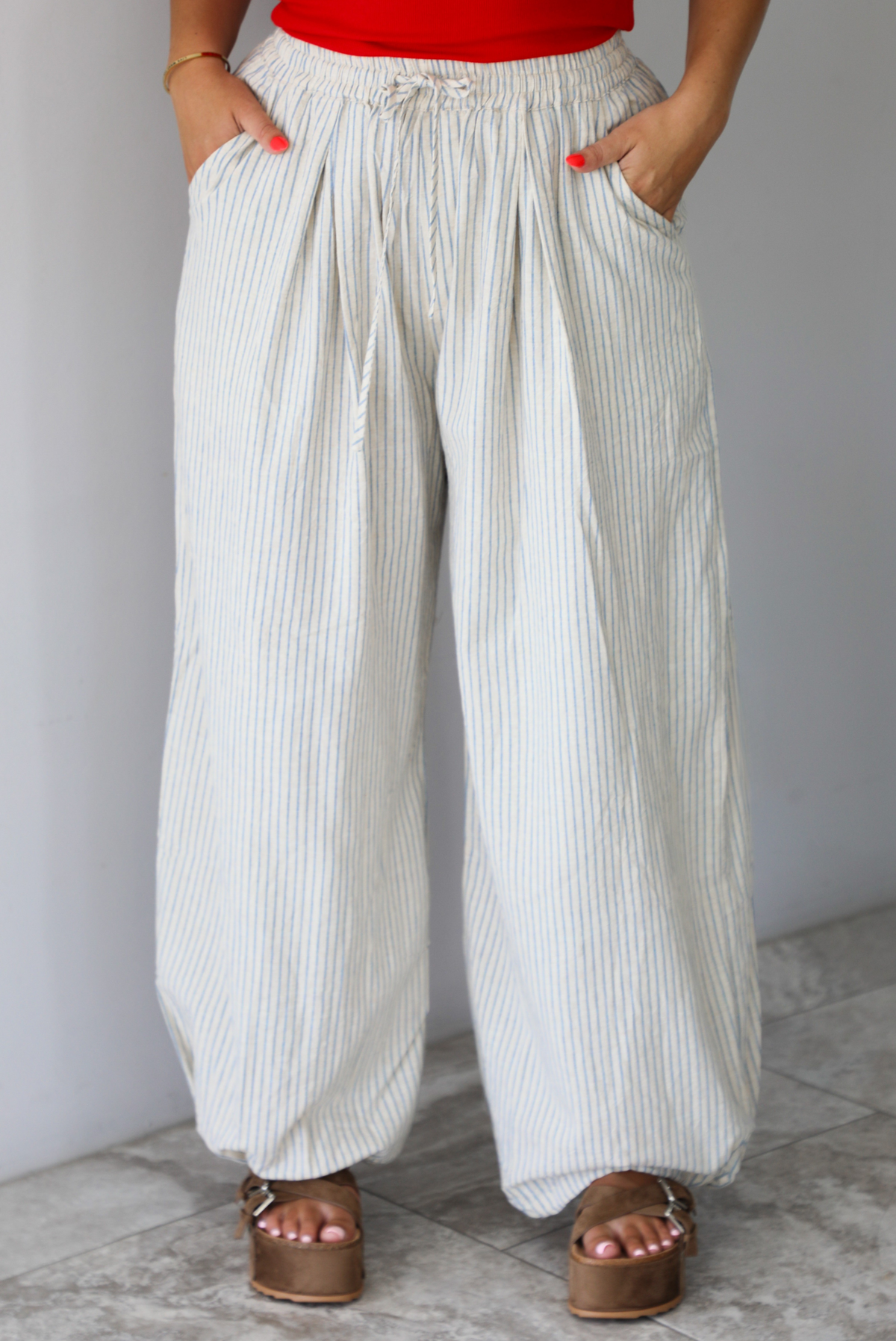 Woman wearing lounge pants that are an ecru color with a blue striped pattern, are high waisted with an elastic and drawstring waistline, and have a slouched design until they cinch at the ankles indoors against a white wall with white marble flooring. The photo shows the models hands in the pockets on either side. 