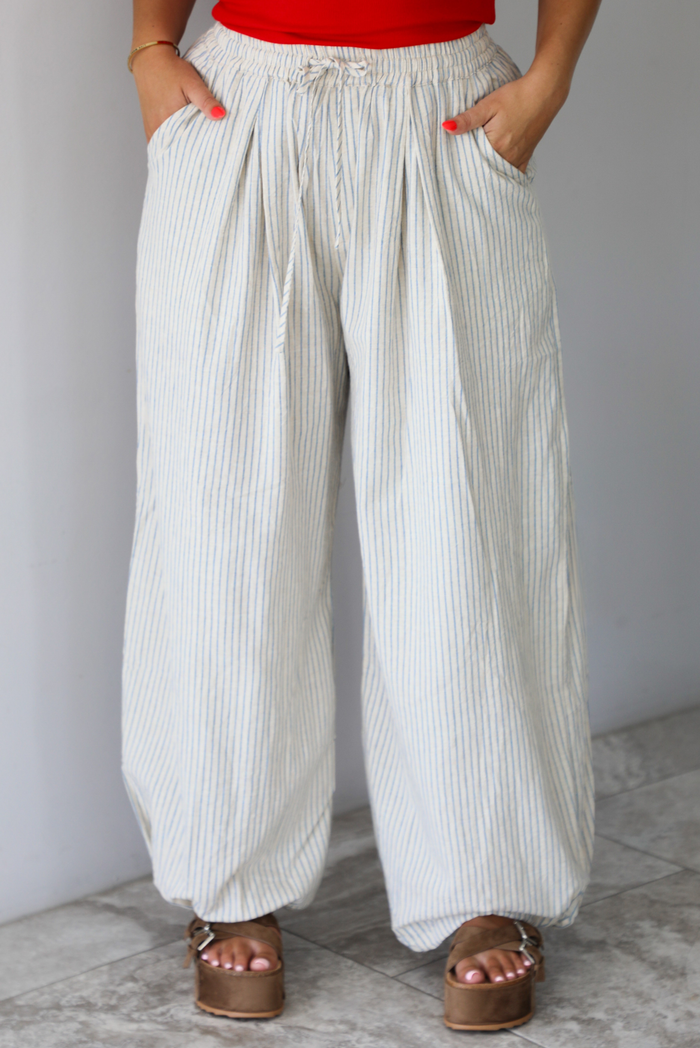 Woman wearing lounge pants that are an ecru color with a blue striped pattern, are high waisted with an elastic and drawstring waistline, and have a slouched design until they cinch at the ankles indoors against a white wall with white marble flooring. The photo shows the models hands in the pockets on either side. 