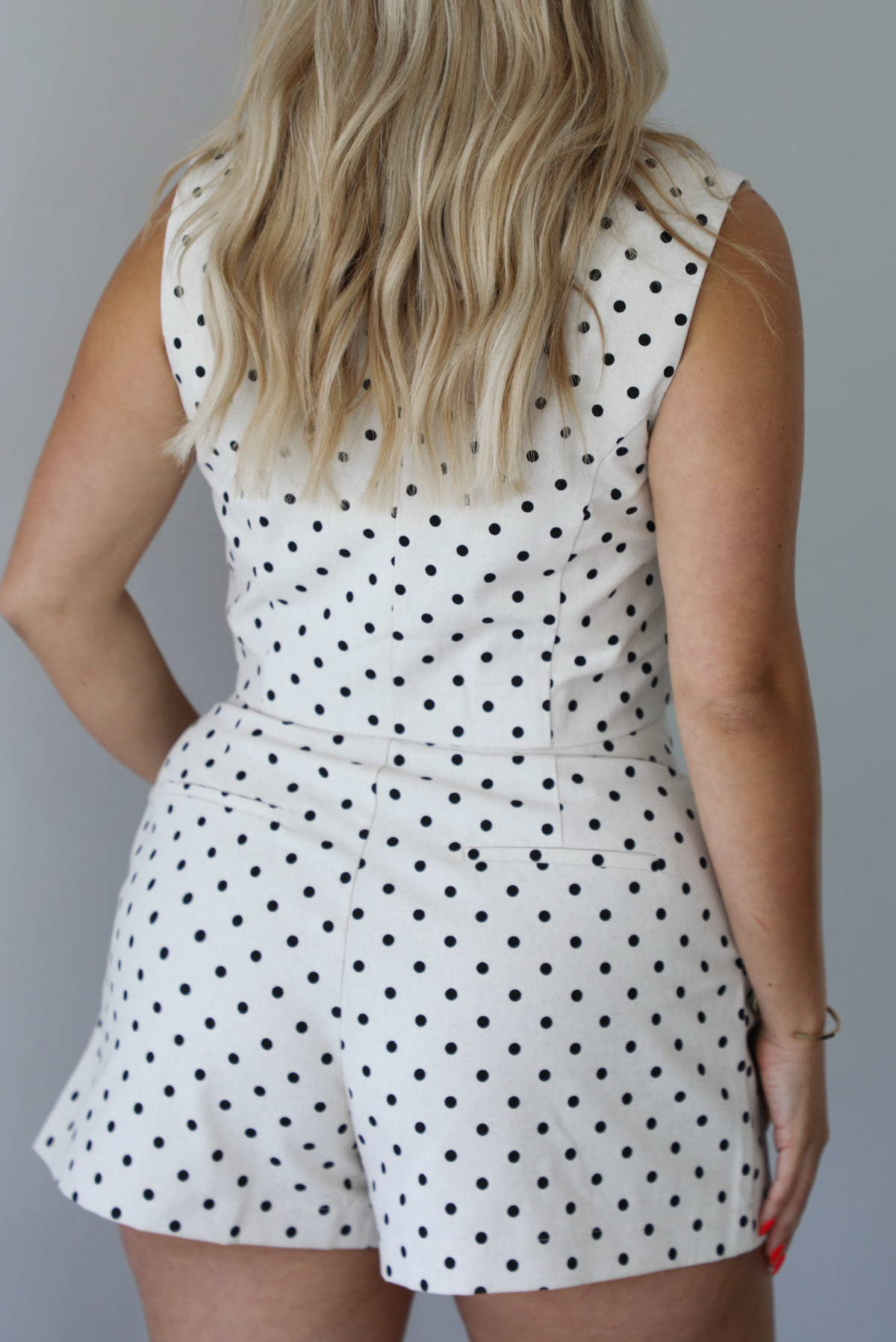 Woman wearing a sand with black polka dots two piece set featuring a cropped top with an asymmetrical cut and high waisted shorts indoors against a white wall. The photo shows a closeup of the back of the set. 