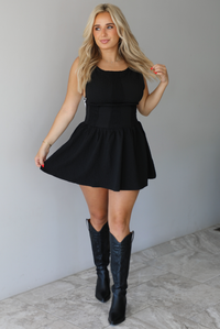 Woman wearing a black mini dress that has a fitted drop waist design and a flowy mini length skirt. The photo is taken indoors against a white wall with white marble flooring. 