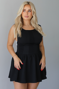 Woman wearing a black mini dress that has a fitted drop waist design and a flowy mini length skirt indoors against a white wall. The photo shows a closeup of the dress, and shows that it has a textured detail on the material.