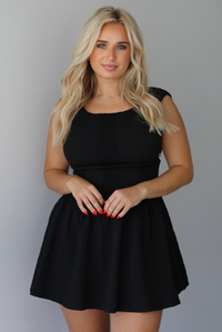 Woman wearing a black mini dress that has a fitted drop waist design and a flowy mini length skirt indoors against a white wall. The photo shows a closeup of the dress, and shows that it has a textured detail on the material.