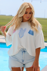 Woman wearing a white oversized box tee with blue and red embroidered graphics on the front with light wash denim shorts while standing next to a pool with greenery in the background. 