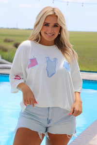 Woman wearing a white oversized box tee with blue and red embroidered graphics on the front with light wash denim shorts while standing next to a pool with greenery in the background. 