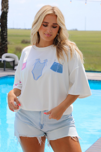 Woman wearing a white oversized box tee with blue and red embroidered graphics on the front with light wash denim shorts while standing next to a pool with greenery in the background. 