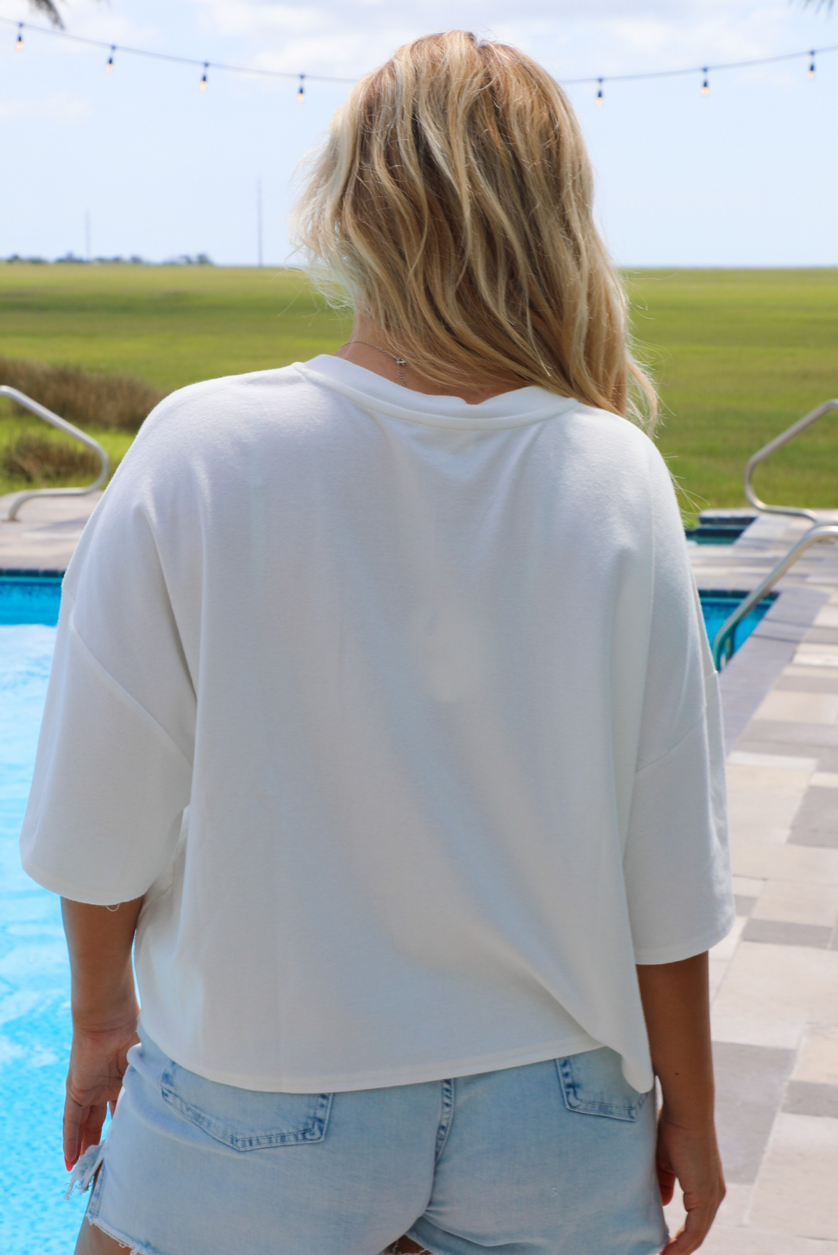 Woman wearing a white oversized box tee with blue and red embroidered graphics on the front with light wash denim shorts while standing next to a pool with greenery in the background.  The photo shows the back of the tee. 