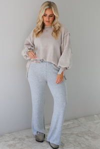 Woman wearing sand colored oversized sweatshirt with exposed seam details and a high-low design with heather grey casual pants. The outfit is styled with grey slip on shoes. The photo is taken indoors against a white wall with white marble flooring. 