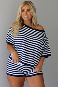 A woman wearing a navy and white horizontally striped off-the-shoulder top and matching striped shorts, standing indoors against a white wall.