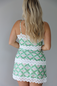 Woman wearing a teal, green, white, and pink dress with white lace trim detailing, thin, spaghetti straps, with a mini length design indoors against a white wall. The photo shows the back of the dress. 