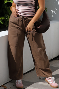 A closeup of a woman wearing a pink, burgundy, and white striped t-shirt, paired with mocha colored barrel denim, along with a brown shoulder bag and pink sneakers, standing outdoors against a low white wall and a stone path. 