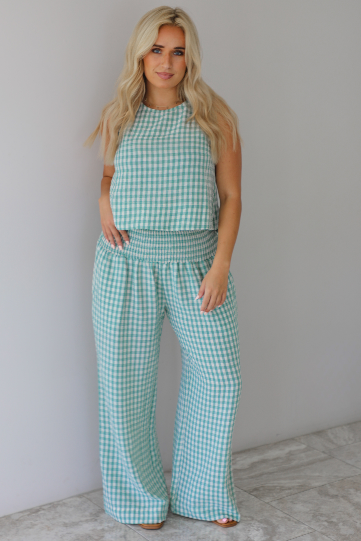 Woman wearing a mint and white gingham set with an oversized top and flared hem pants indoors against a white wall with white marble flooring. 