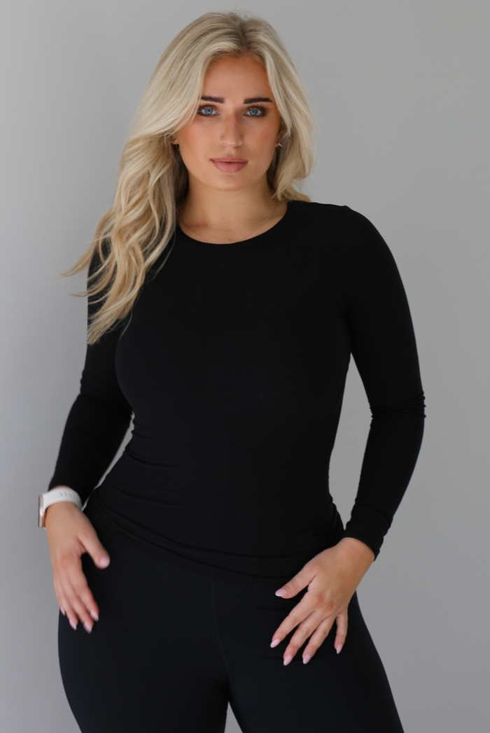 Woman wearing a black, high neckline, long sleeved fitted top that has a ribbed design to the material. It's paired with black leggings. The photo is taken against a white wall. 