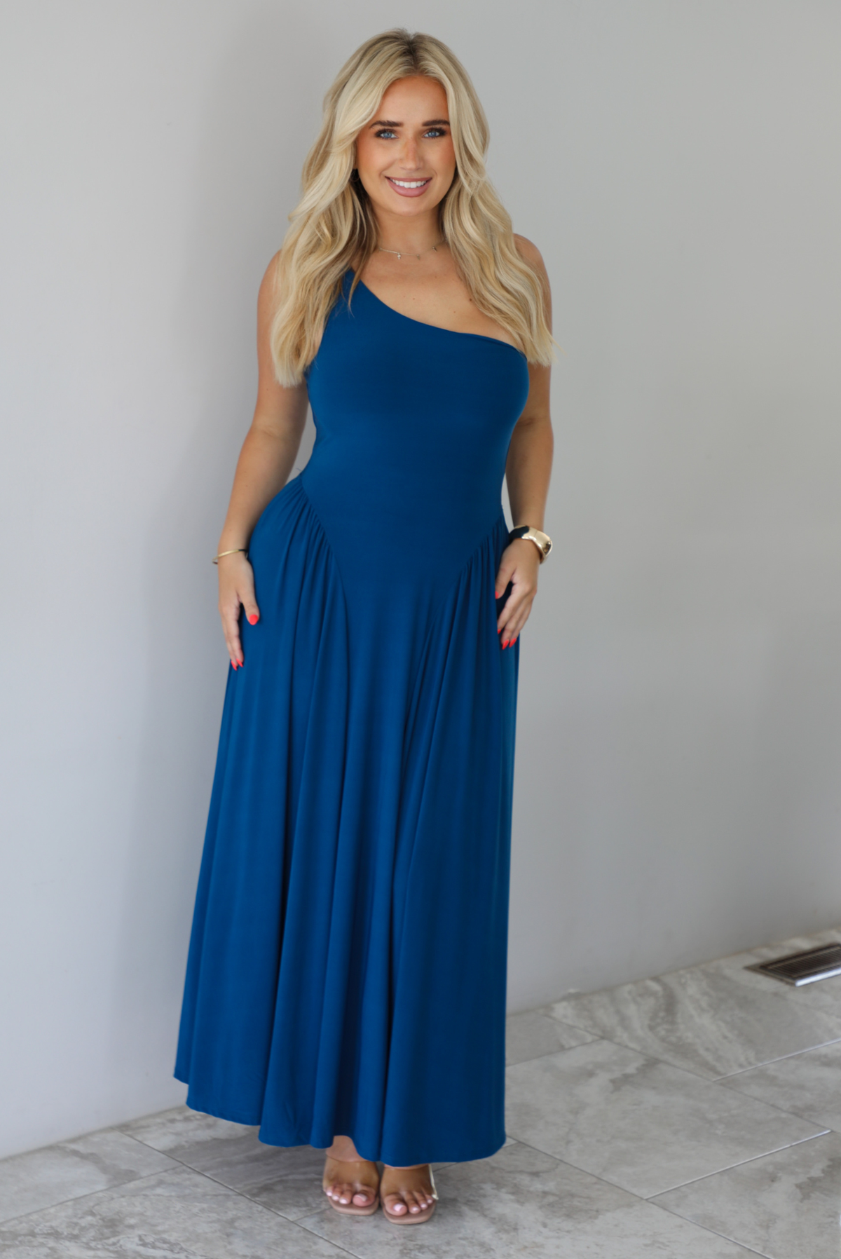 Woman wearing a blue one strap maxi dress indoors against a white wall with white marble flooring. 
