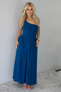 Woman wearing a blue one strap maxi dress indoors against a white wall with white marble flooring. 