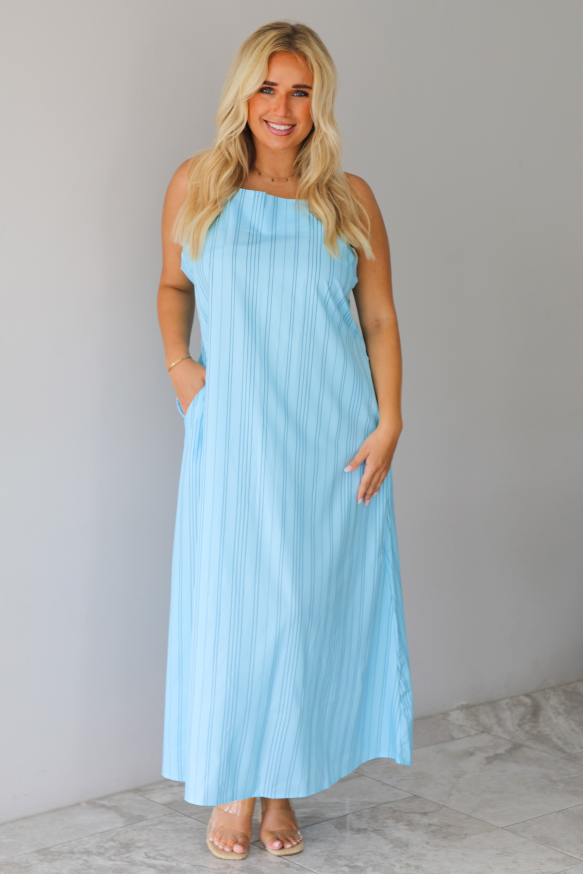 A woman modeling a light blue sleeveless maxi dress with vertical grey pinstripes and side pockets, standing indoors against a white wall with white marble flooring. 