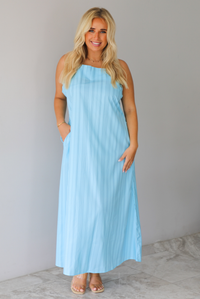 A woman modeling a light blue sleeveless maxi dress with vertical grey pinstripes and side pockets, standing indoors against a white wall with white marble flooring. 