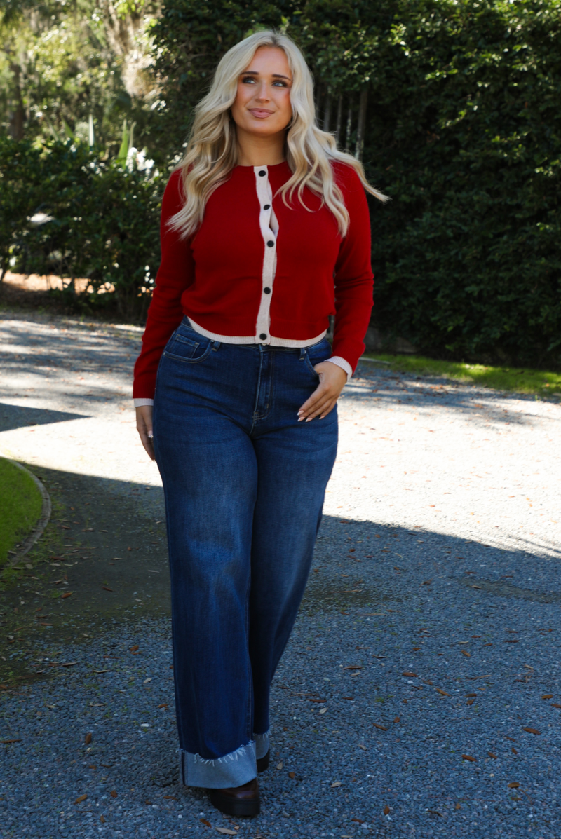 Woman model wearing a red cardigan with sand trim details and black button closures in the front with dark wash denim. The photo is taken outdoors on gravel with greenery in the back. 