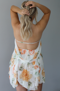 Woman wearing a white, strapless, babydoll cut dress that has an orange and green floral print pattern and flowy skirt indoors against a white wall. The photo shows the back of the dress, which is mainly backless with an elastic band around the back to hold the dress up with a zipper closure and drape detail. 