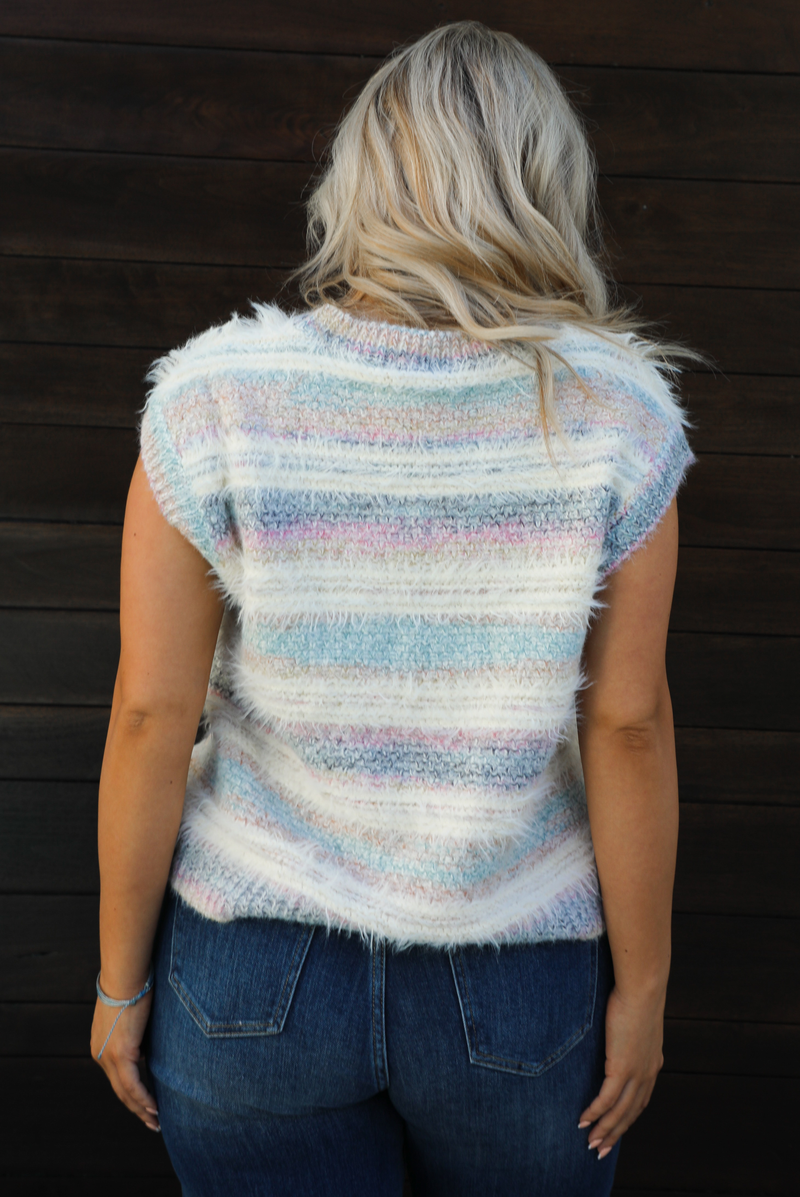 Woman model wearing a soft, striped sweater vest with dark wash denim jeans. The photo showcases a closeup of the back of the sweater vest. The photo is taken outdoors against a dark brown wooden door. 