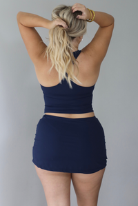 A woman wearing a navy two piece matching athletic set featuring a halter tank top and fitted mini skirt, standing indoors against a white wall. The photo shows the back of the set. 