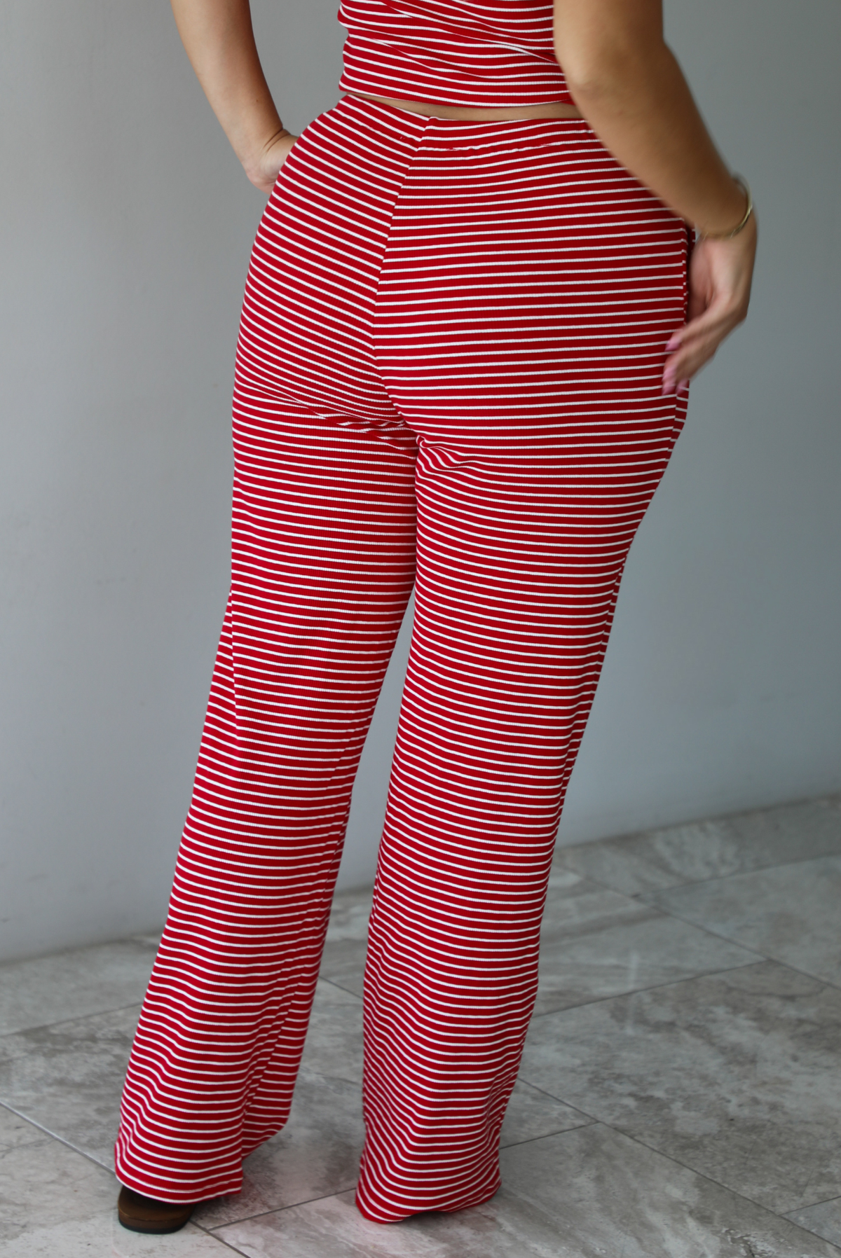 A woman modeling a red and white striped sleeveless set consisting of a tank top with non-functional button closures in the front and high waisted pants with a drawstring waistline and pockets on either side indoors against a white wall with white marble flooring. The photo shows a closeup of the back of the pants. 
