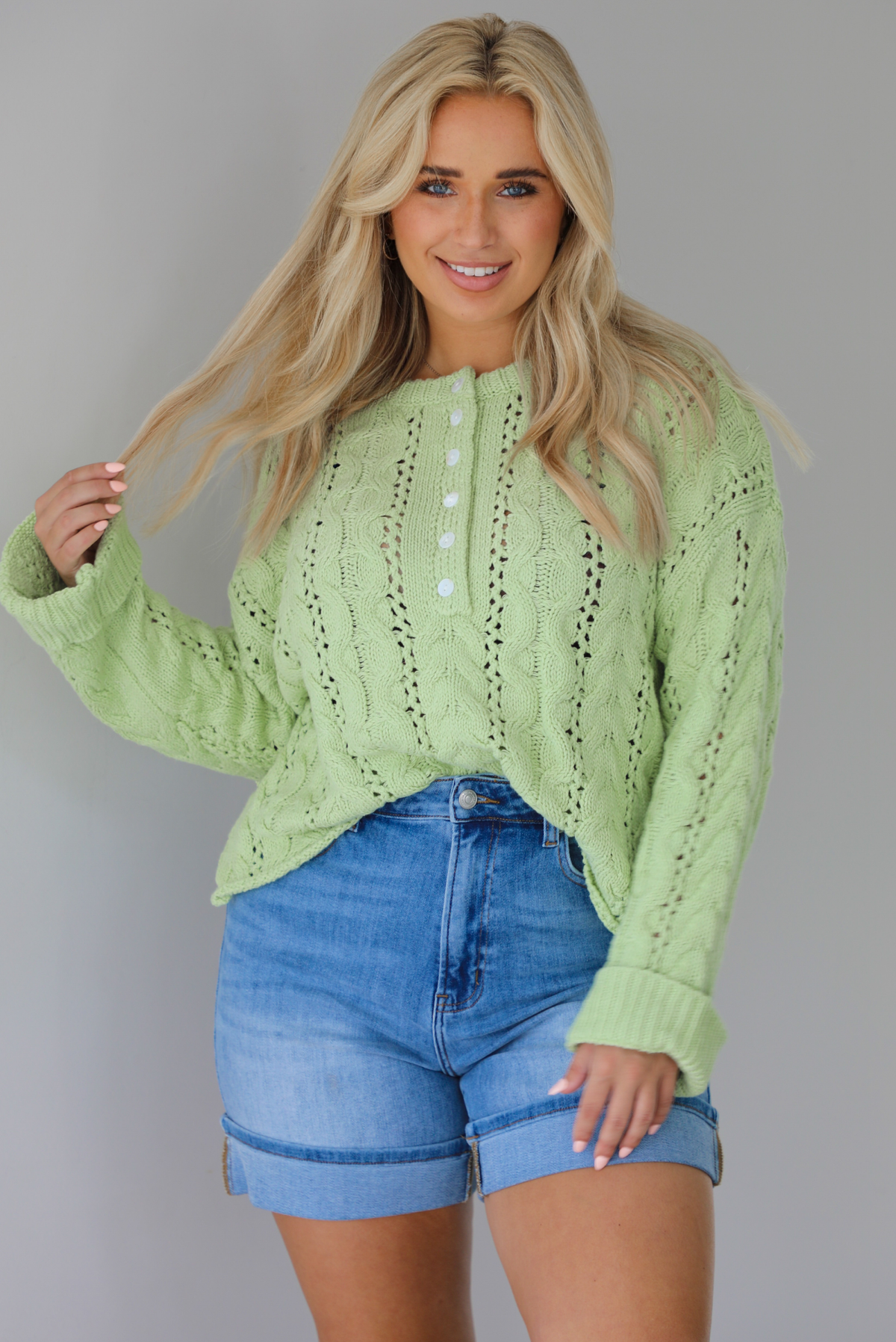 Woman wearing a green, long sleeved sweater with a loose knit design, functional button closures in the front, cuffed sleeves, and a raw, unfinished hem with medium wash denim shorts indoors against a white wall.