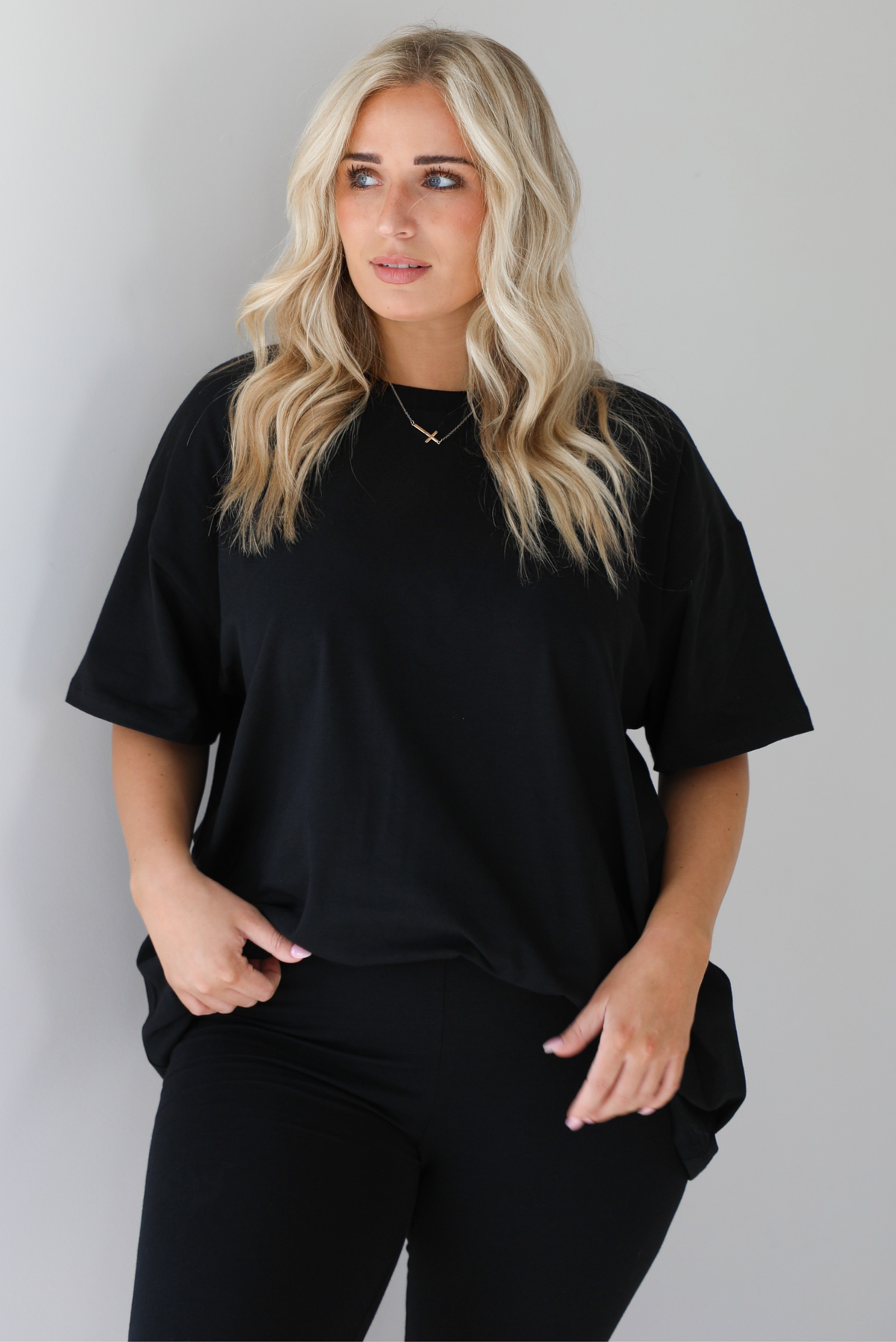 Woman wearing a black two piece set featuring an oversized t-shirt and fitted leggings. The photo shows a closeup of the oversized top. The photo is taken indoors against a white wall.