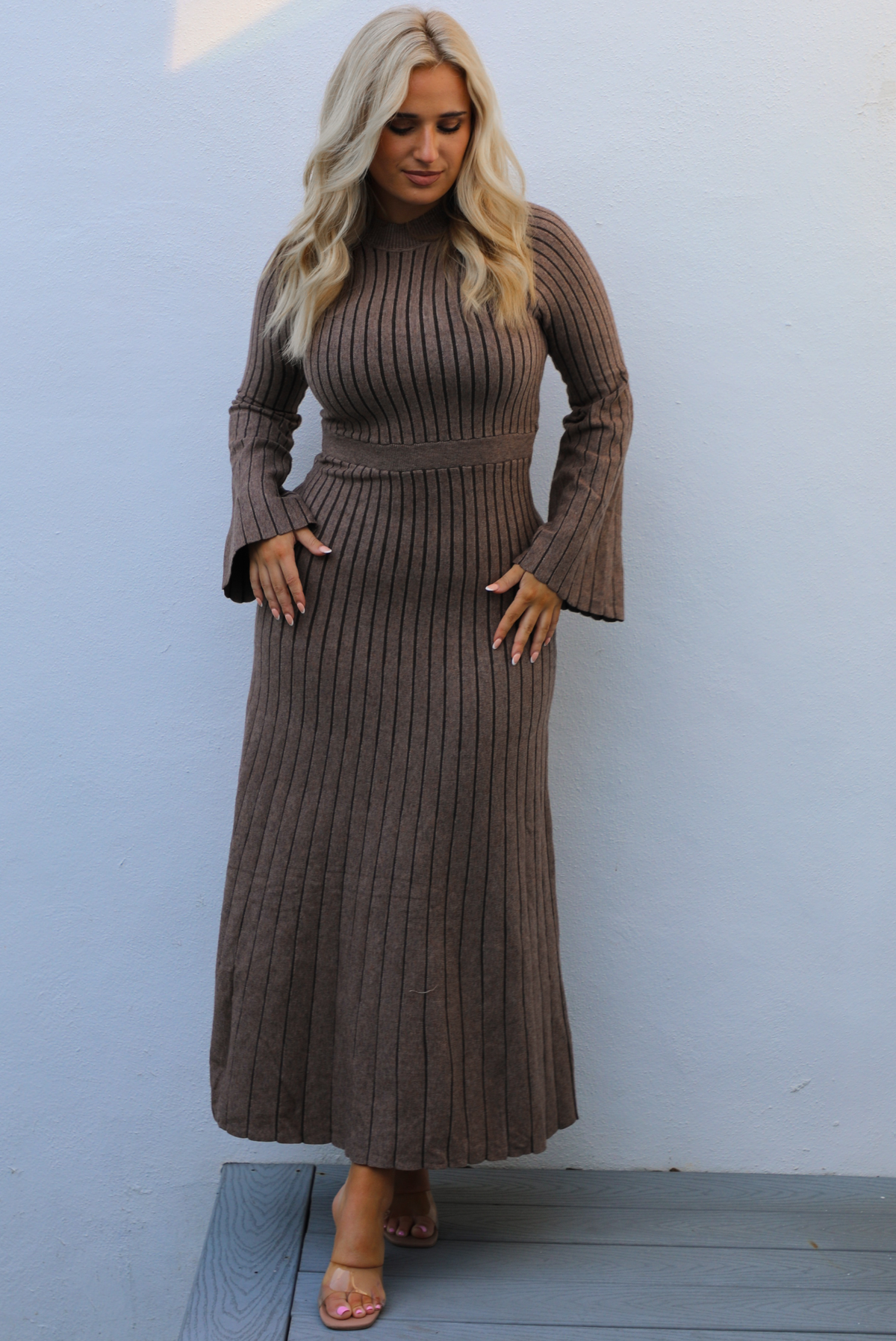 Woman wearing a taupe colored dress that has a vertical ribbed pattern throughout, a high neckline, fitted bodice, cinched waistline, long sleeves that flare slightly, and a maxi length skirt that flows at the bottom. The photo is taken outdoors against a white wall. 