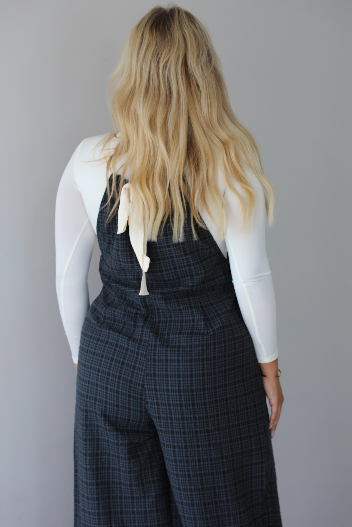 A woman shown from behind modeling a white long-sleeve top and dark gray plaid wide-leg overalls, standing indoors against a white wall.