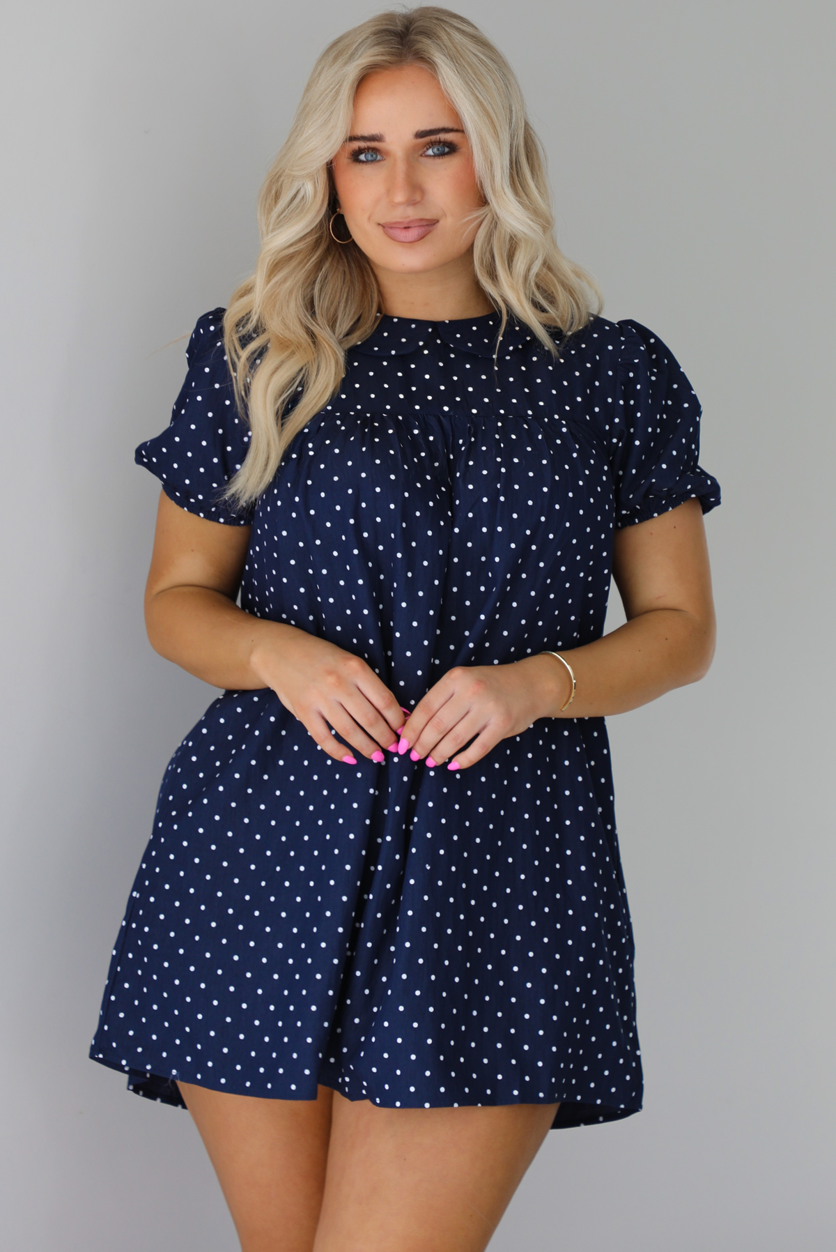 Woman wearing a navy baby-doll cut dress with a white polkadot print pattern throughout. The dress has puffed short sleeves and functional pockets on either side. The photo is taken indoors against a white wall.