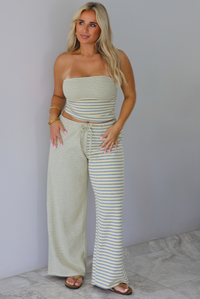 A woman wearing a pale yellow and heather grey striped two piece set with a sleeveless tube top and wide-leg pants, standing indoors against a white wall with white marble flooring. 