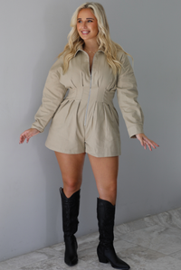 A woman wearing a sand colored long sleeved romper with a collared neckline, functional zipper closure in the front, pleated details throughout, functional pockets on either side, paired with black colored knee-high boots, standing indoors against a white wall with white marble flooring. 