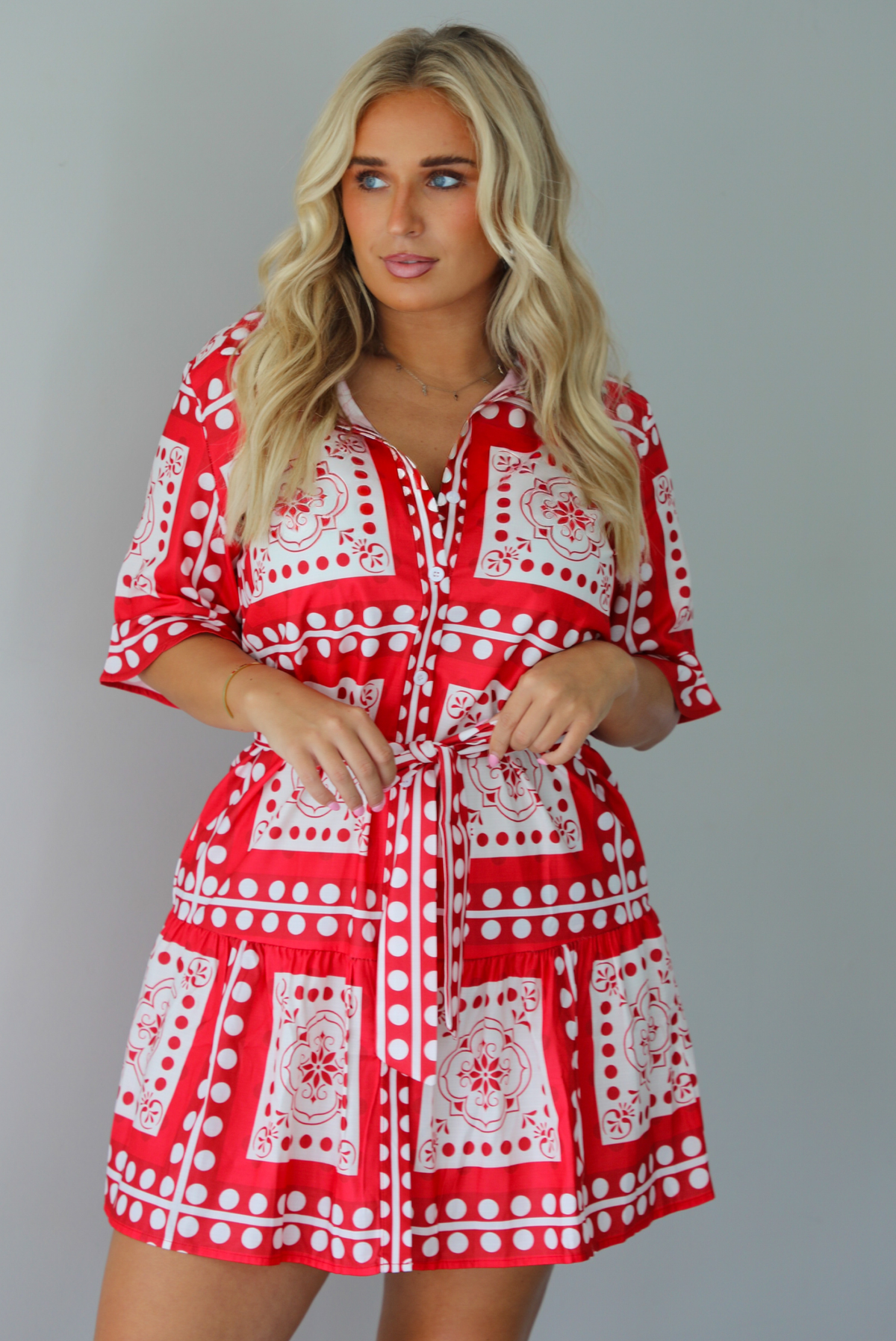 A woman modeling a red and white patterned short-sleeve mini dress with button closures along the front and a tie waist, standing indoors against a white wall.