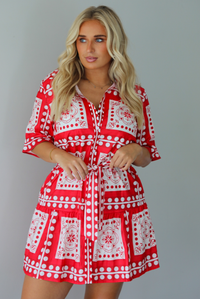 A woman modeling a red and white patterned short-sleeve mini dress with button closures along the front and a tie waist, standing indoors against a white wall.