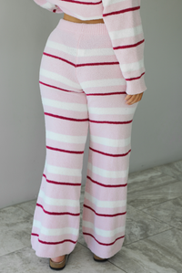 Woman wearing a pink set with a darker pink and white striped design. The set consists of an oversized sweater top and high waisted pants. The photo shows a closeup of the back of the bottoms. The photo is taken indoors against a white wall with white marble flooring. 