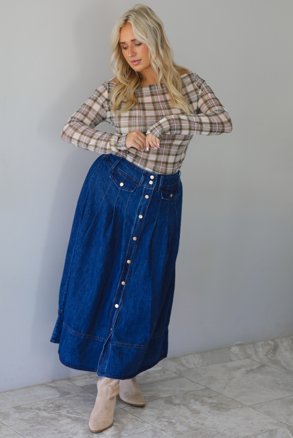 A woman wearing a beige and brown plaid long-sleeve top and a high-waisted blue denim button-front midi skirt, standing indoors against a white wall with white marble flooring. 